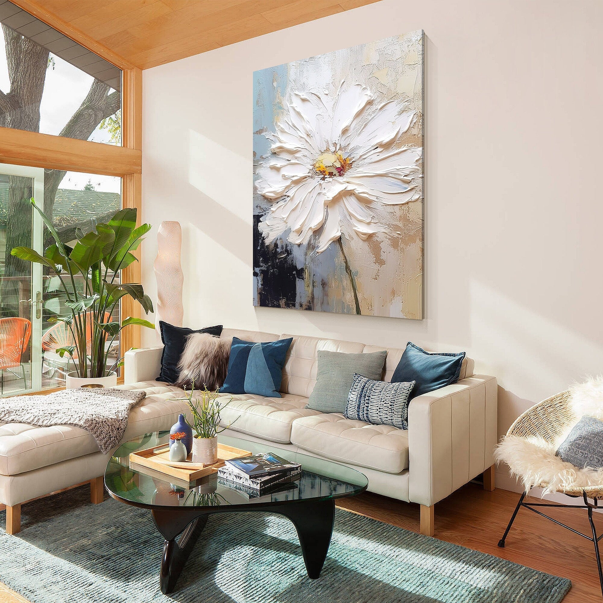 A white canvas painting of a large white flower with a yellow center. The painting is hanging on a white wall above a white couch with blue pillows.